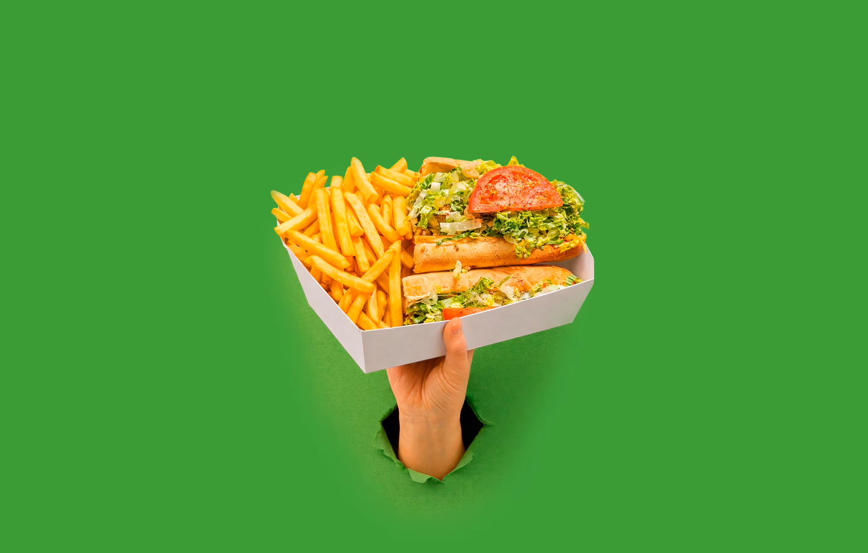 Hand holding a white tray with French fries and a sandwich filled with lettuce and tomato against a green background.